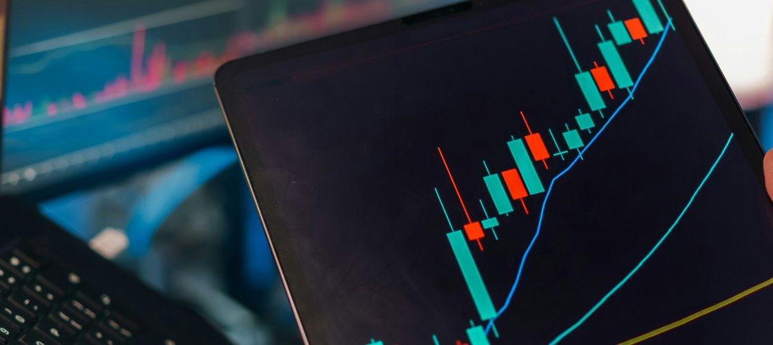 A tablet showing a candlestick chart held in front of a trading monitor and laptop