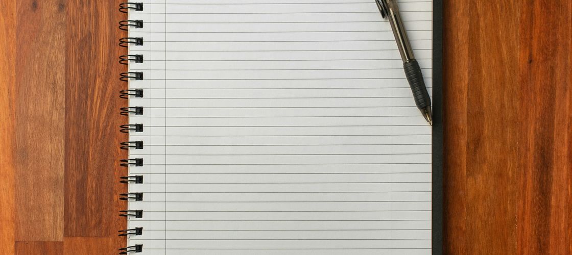 An open notebook and pen on a wooden desk beside a laptop